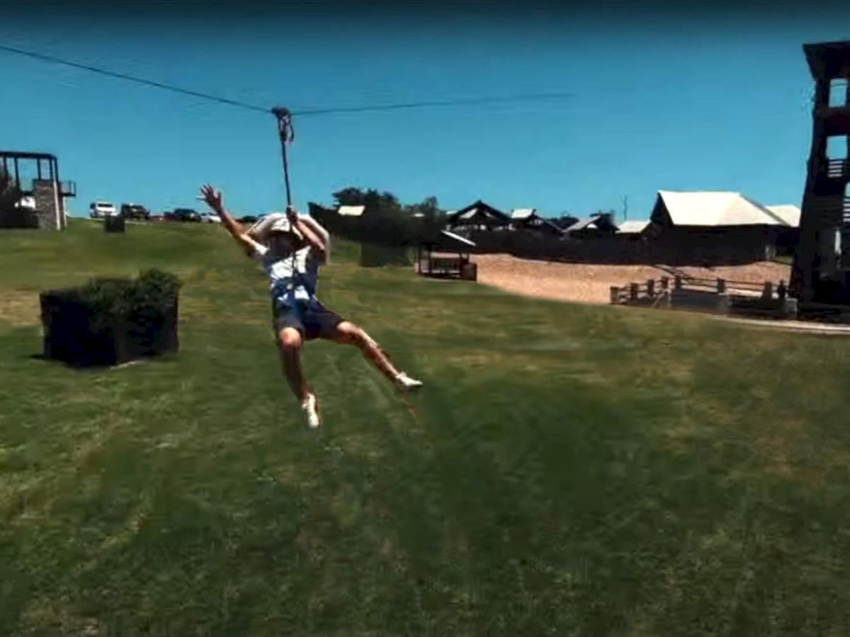 A person is zip-lining across a grassy field with a clear blue sky and some playground structures in the background.