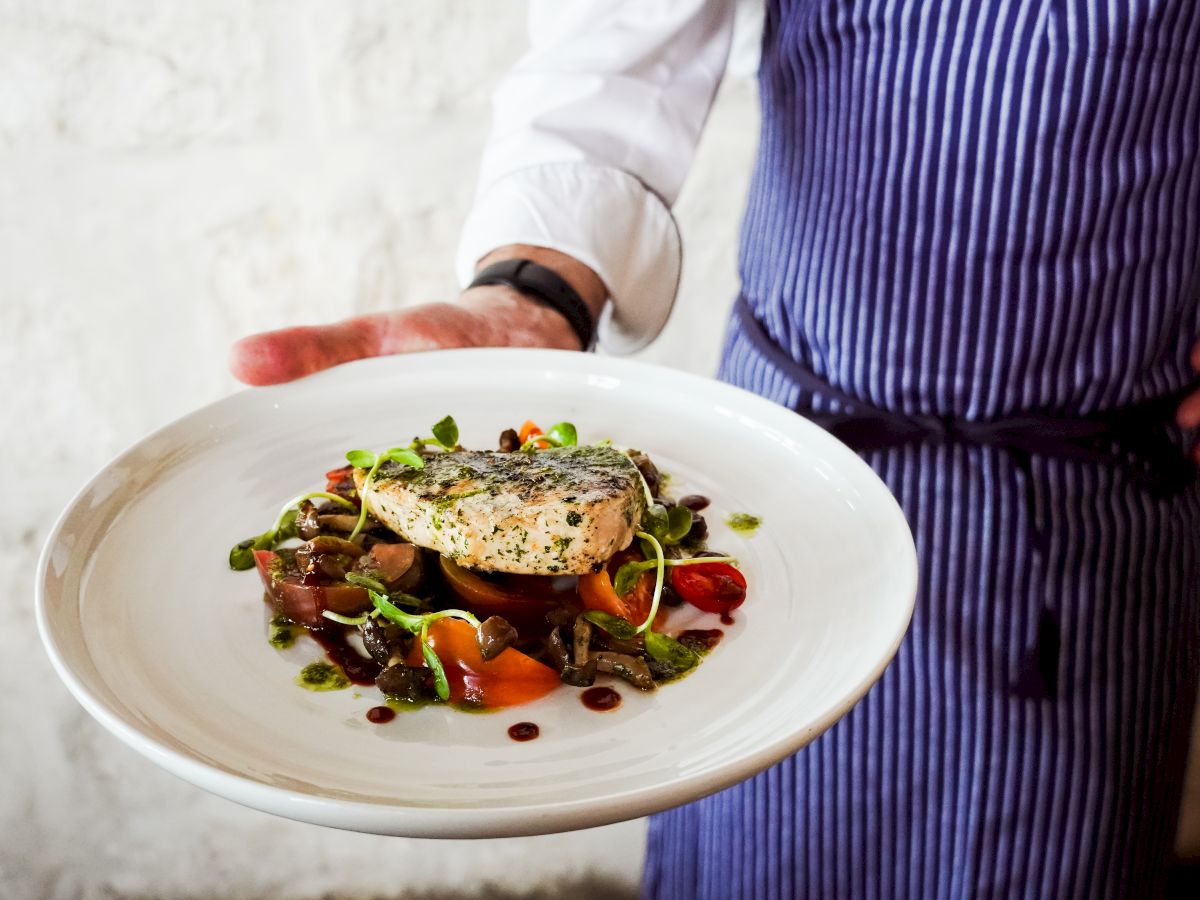 A chef in a striped apron presents a plated, colorful dish with greens, olives, and a seared fish fillet on a white plate.
