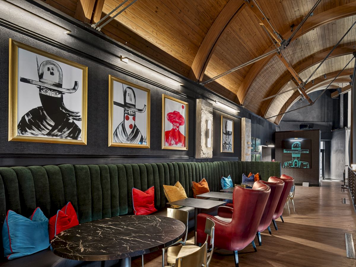 A stylish lounge with a long black tufted sofa, colorful chairs, round marble tables, and framed black-and-white portraits along a wood-arched ceiling, elegant vibe.