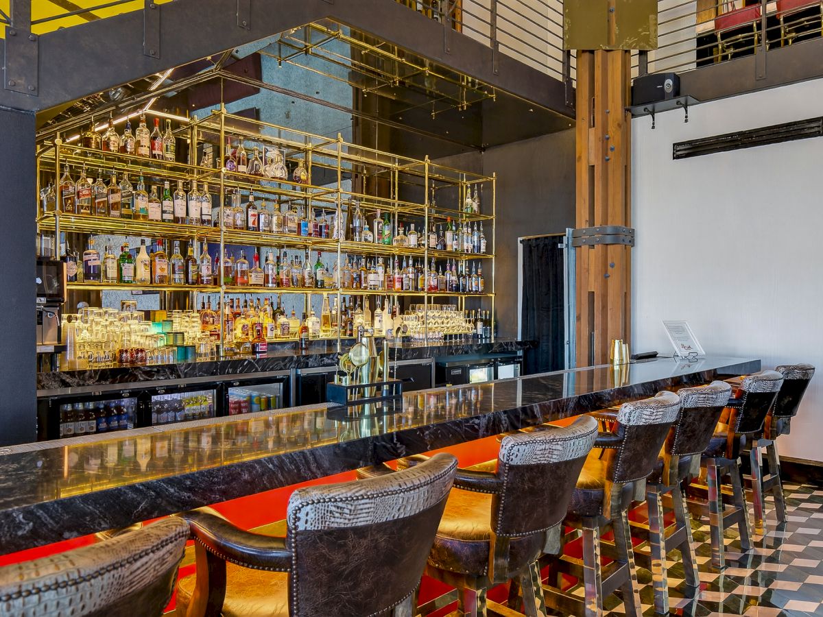 A stylish bar with a long counter, row of high chairs, and shelves stocked with bottles in the background, under warm lighting.